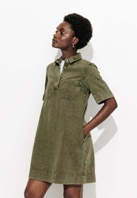 Robe à manches courtes, couleur vert kaki, en tissu doux, avec un col, une patte de boutonnage et deux poches avant.