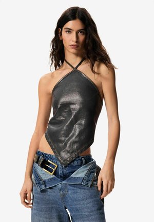 Jeune femme portant un haut halter sans manches, noir métallisé avec un ourlet angulaire, associé à un jean bleu et une large ceinture noire avec boucle dorée.