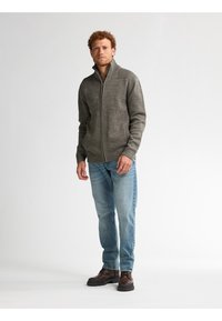 Grauer Reißverschluss-Pullover mit strukturierten Schulterakzenten, getragen über einem beigen Oberteil. Kombiniert mit hellblauen Jeans und dunkelbraunen Schuhen.