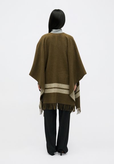 Poncho verde-azeitona com riscas cinzas e barra com franjas, feito de tecido suave. Modelo veste calças pretas e um suéter cinza de gola alta.