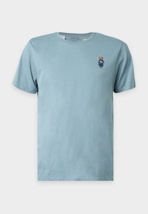 Hellblaues T-Shirt mit kurzen Ärmeln und Rundhalsausschnitt mit einem kleinen bestickten Teddybär mit blauer Jacke auf der linken Brust.