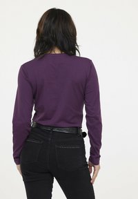 T-shirt à manches longues violet avec col rond. Associé à un jean noir taille haute, orné de détails cloutés sur les poches arrière.