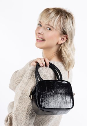 Borsa a tracolla nera in patinato con forma quadrata, texture liscia e doppi manici. Chiusura con cerniera e leggera lucentezza. Indossata sopra un maglione chiaro.