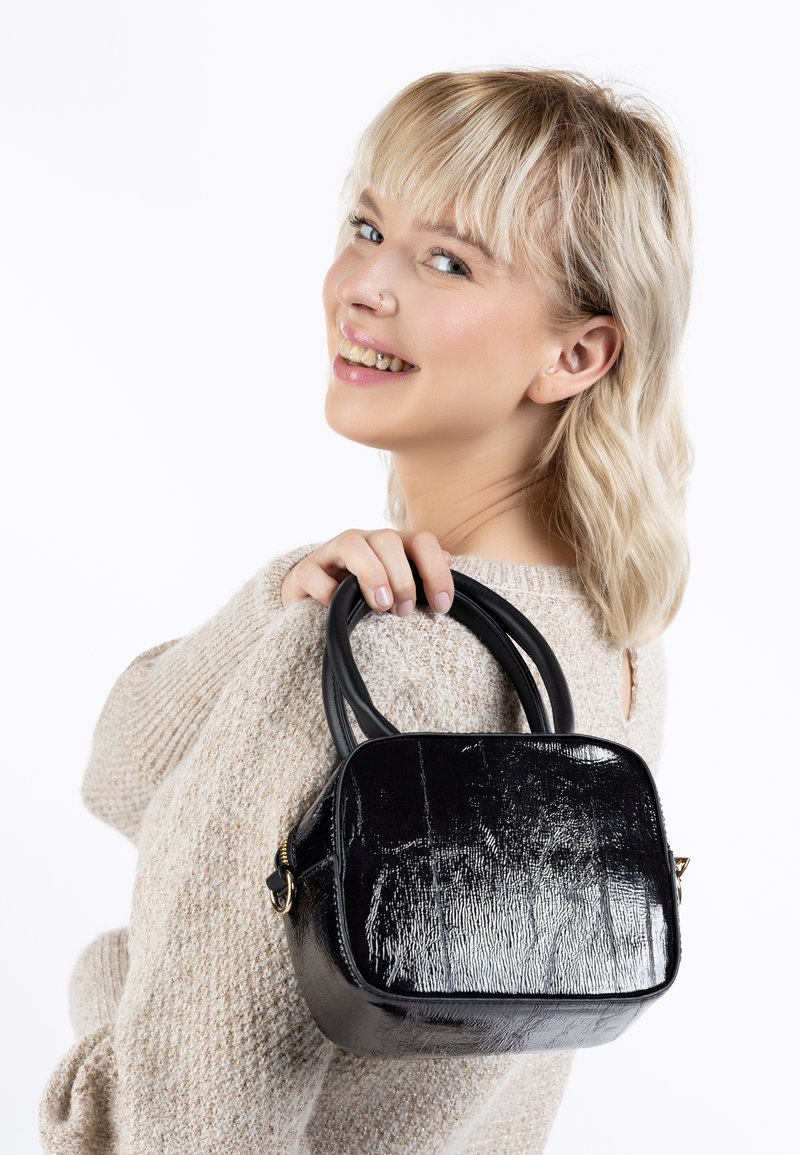 Borsa a tracolla nera in patinato con forma quadrata, texture liscia e doppi manici. Chiusura con cerniera e leggera lucentezza. Indossata sopra un maglione chiaro.