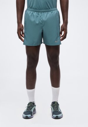 Man draagt turquoise sportshorts, witte enkelsokken en zwarte en turquoise hardloopschoenen, staand tegen een effen witte achtergrond.