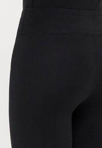 Artimiausias plano kadras asmens, vilkinčio aukšto juosmens juodas leggins, kuriuose matomi siūlės prie juostos ir šoninio siūlo.