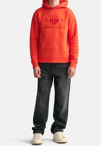 Rote Kapuzenjacke mit eingeprägtem Logo, kombiniert mit weit geschnittenen dunkelgrauen Jeans und weißen Sneakers. Die glatte Stoffstruktur ist sichtbar.