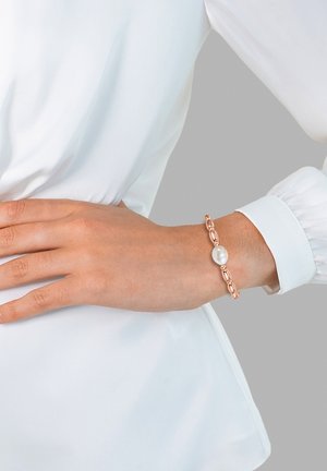Pulsera de oro rosado con eslabones lisos y un acento de perla central, llevada en una muñeca sobre una blusa blanca.