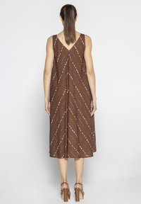Brąsowa sukienka bez rękawów z białymi ukośnymi paskami, z dekoltem w kształcie litery V, do kolan, wykonana z teksturowanej tkaniny; noszona z brązowymi sandałami na obcasie.