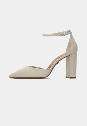 Kengät korkea korko teräväkärkinen beige, nilkkahihnalla ja kullanvärisellä soljella, paksu korko ja sileä matta pinta.