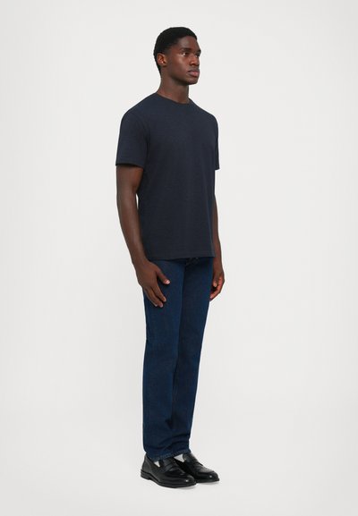Young man standing, wearing a plain dark navy t-shirt, dark blue jeans, and black leather loafers against a white background.