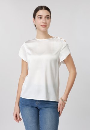 Mladá žena v bielej saténovej blúzke s krátkym rukávom a gombíkmi na pleciach, oblečená v modrých džínsoch a so zlatým náramkom, pózujúca na jednoduchom sivom pozadí.