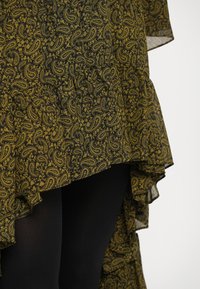 Robe à motif paisley noir et jaune avec ourlet volanté, design superposé et texture transparente, portée sur des leggings noirs.