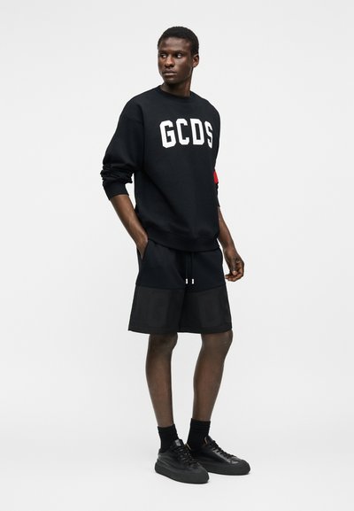 Άνδρας στέκεται με τα χέρια στις τσέπες, φορώντας μαύρο φούτερ GCDS, μαύρα σορτς, μαύρες κάλτσες και μαύρα αθλητικά παπούτσια σε λευκό φόντο.