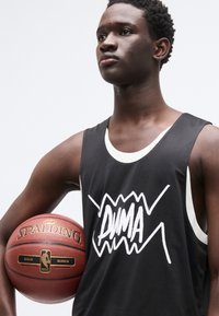 Μαύρο αμάνικο μπλουζάκι μπάσκετ με λευκό λογότυπο και λεπτομέρειες, με λαιμόκοψη σε αντίθετο χρώμα. Το μοντέλο κρατάει μια κόκκινη και καφέ μπάλα μπάσκετ Spalding.