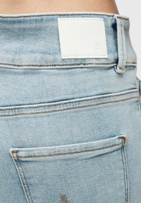 Κοντινό πλάνο γαλάζιου τζιν υφάσματος που δείχνει την πίσω τσέπη, τη θηλιά της ζώνης και ένα λευκό ορθογώνιο μπαλώμα κοντά στη μέση.