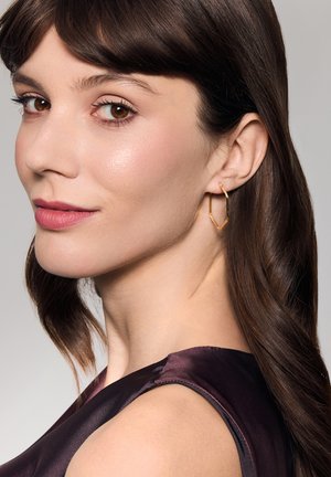 Złote geometryczne kolczyki w kształcie koła prezentowane na modelce z ciemnymi brązowymi włosami. Kolczyki mają wypolerowane, metaliczne wykończenie i kanciasty design.