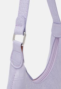 Lila handväska med en texturerad imitation läderdesign, en böjd form och silverfärgad hårdvara. Innehåller ett zip-fack och justerbar rem.