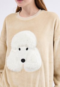 Beżowa, pluszowa bluza z nadrukiem białego pudla, o puszystej teksturze, okrągłej głowie i czarnych oczach, na tle miękkim i gładkim.