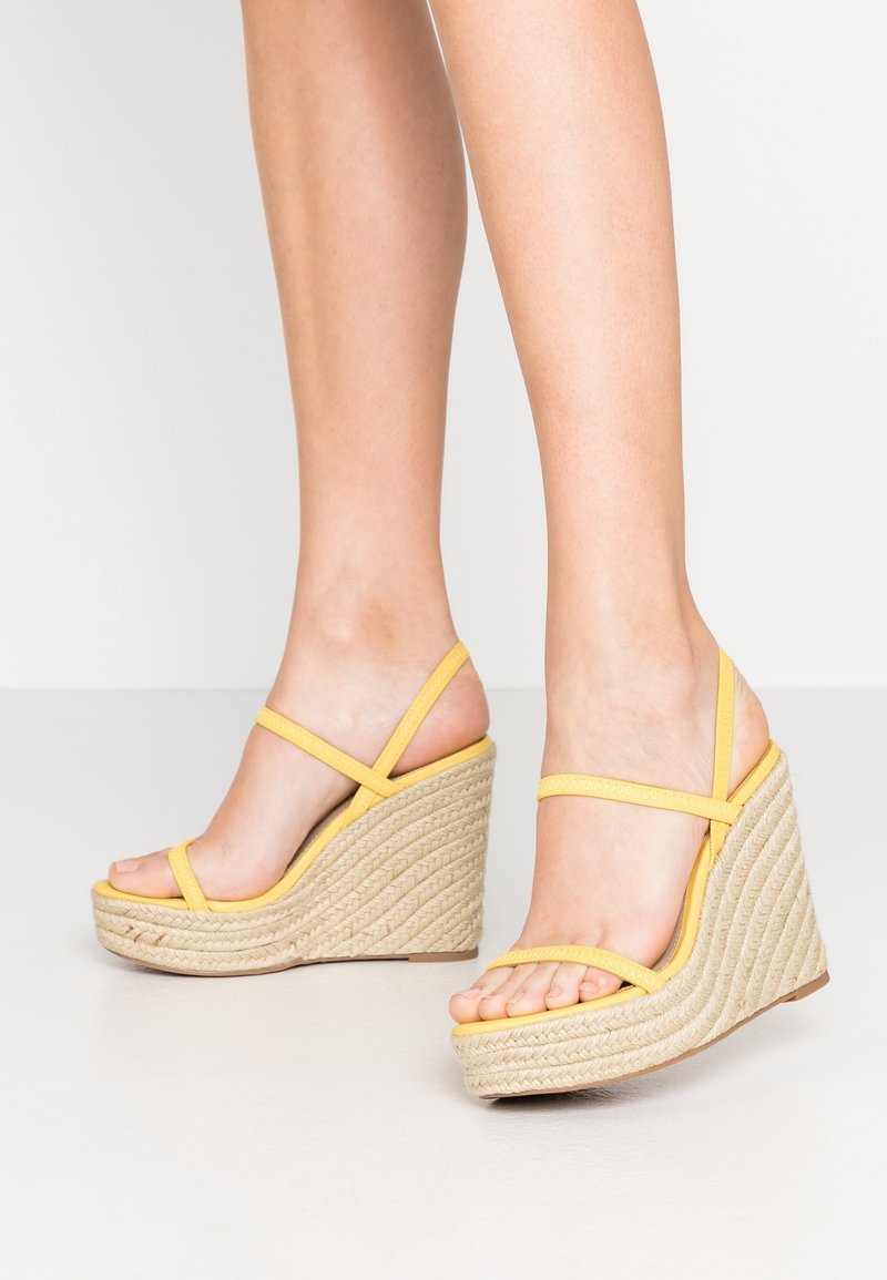 Jambes portant des sandales compensées jaunes à brides avec des talons plateformes en corde tressée, debout sur un sol clair contre un fond blanc.