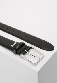 Cinturón de cuero negro con una hebilla rectangular plateada, que presenta un logo de la marca. El cinturón tiene varios agujeros y una textura suave.