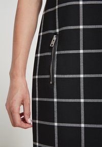 Robe noire avec un motif à carreaux blancs, présentant un détail de poche zippée visible sur le côté. Tissu lisse avec une coupe ajustée.