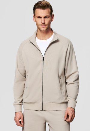 Mężczyzna w jasnobeżowej kurtce na zamek, nałożonej na białą koszulę, stoi na tle jednolitego szarego tła.