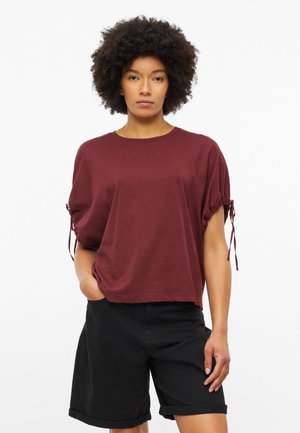 Vrouw met krullend haar die een losse bordeauxrode top met korte mouwen en zwarte knielange shorts draagt tegen een effen witte achtergrond.