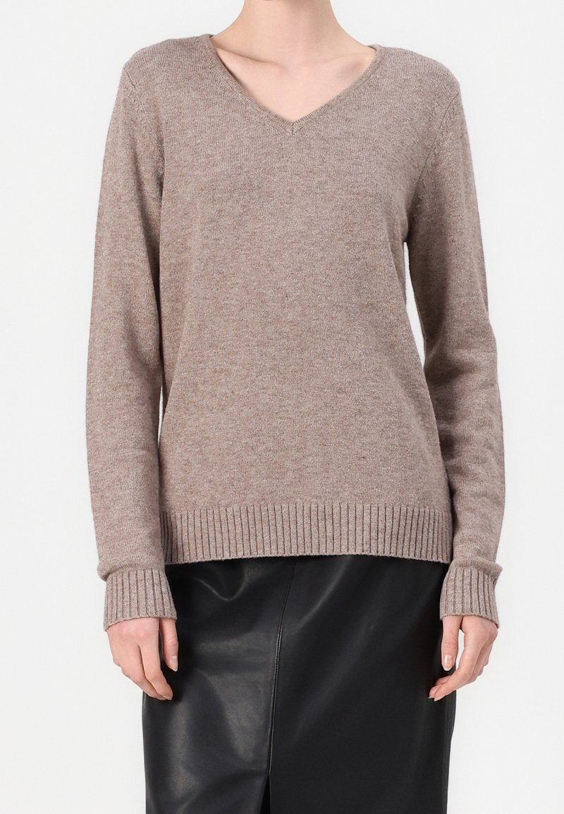 Personne portant un pull taupe à manches longues et col en V ainsi qu'une jupe en cuir noire, debout devant un fond clair uni.