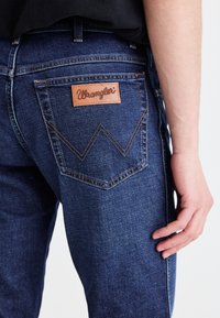 Tamsiai mėlynos džinsinės kelnės su odiniu Wrangler ženklu. Yra matomas siuvinėjimas ir išskirtinis „W“ formos kišenių dizainas.