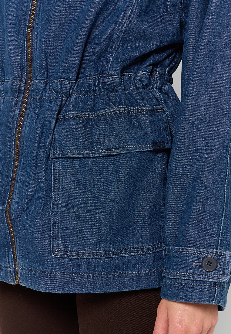 Chaqueta de mezclilla con cremallera frontal, cintura elástica, gran bolsillo con solapa, puño abotonado, llevada sobre pantalones oscuros.