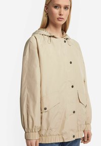 Beige huva jacka gjord av lätt material, med elastiska ärmslut, framknappning och två stora sidofickor.