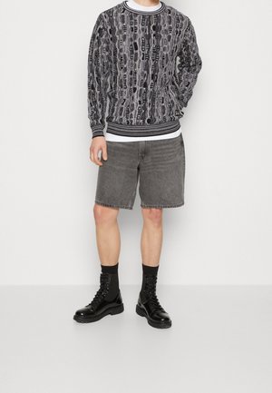 Pull en noir et blanc avec un motif complexe, col rond à côtes, associé à un short en jean gris, des bottes noires à lacets et des chaussettes.