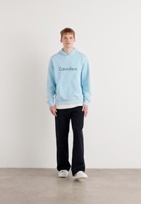 Svetlo modrá mikina s kapucňou s logom "Calvin Klein", predná klokaní vrecko, vrstvená na bielom tričku, v kombinácii s tmavými nohavicami a bielymi topánkami.