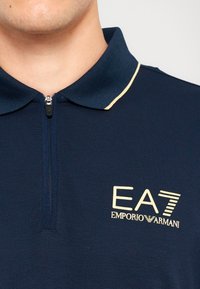 Jūreivio polo marškiniai su užtrauktuku, tekstūruotu apykaklės kraštu su auksine apdaila ir priekinis logotipas "EA7 Emporio Armani" auksinės spalvos. Lygus paviršius.