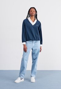 Pullover azul-marinho de mangas longas com colarinho e punhos brancos, combinado com jeans azul-claro e ténis brancos com um logotipo azul-marinho.