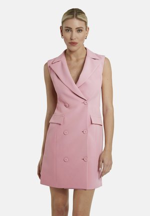 Frau mit blondem Haar in einem ärmellosen, zweireihigen rosa Blazer-Kleid mit Revers und Klappentaschen, stehend vor einem schlichten weißen Hintergrund.