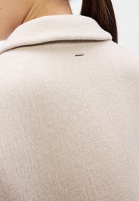Ελαφρώς μπεζ υφασμάτινο πουλόβερ με υφή, με γιακά και μικρό μεταλλικό ορθογώνιο διακοσμητικό στο πίσω μέρος.