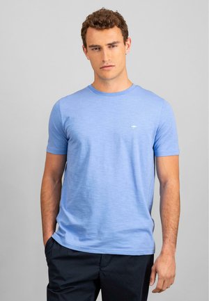 T-shirt bleu clair à manches courtes en tissu doux, avec un col rond et un petit logo sur la poitrine. Porté avec un pantalon sombre.