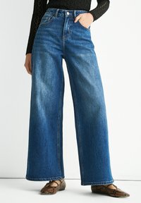 Wide-leg denim jeans in medium blue with a high waist. Features five pockets, belt loops, and a seamless hem. Styled with patterned shoes.