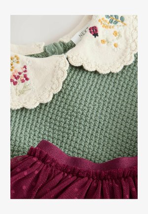 Vihreä neulepusero, jossa on kerroksellinen kreemivärinen kaulus, johon on brodattu kukkakuviota. Puseroa on yhdistetty tummanpunaisesta tulitöyhdöstä valmistettuun hameeseen, jossa on pilkullinen pinta.