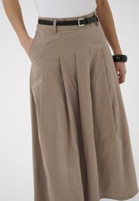 Pantaloni bej cu picioare largi și pliuri, curea neagră și top alb; mâna stângă a persoanei este în buzunar, iar încheietura dreaptă poartă o brățară neagră.