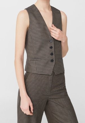 Brązowa, bezrękawnikowa kamizelka w kratę z czterema guzikami, zestawiona z pasującymi, wysokim stanem, szerokimi spodniami na jasnym tle.