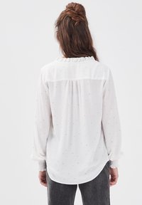 Femme avec de longs cheveux bruns en demi-queue, portant un blouse blanche à manches longues avec un motif subtil et un pantalon gris foncé, vue de dos.
