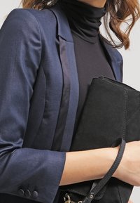Granatowa marynarka z satynowym klapkiem, noszona na czarnym golfie. Trzyma czarną zamszową kopertówkę z paskiem i detalami.