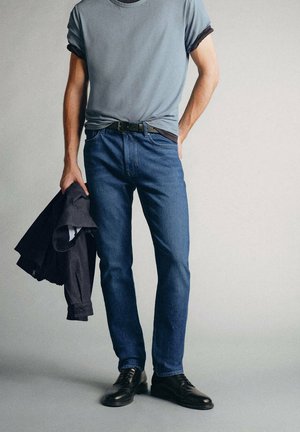 Homme portant un jean bleu, un t-shirt gris, une ceinture noire et des chaussures habillées noires, tenant une veste sombre dans une main devant un fond uni.