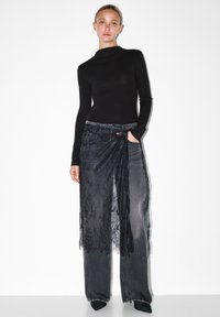 Musta pitkähihainen paita, joka on yhdistetty leveisiin tummansinisistä denimihaalareihin, joissa on läpikuultava musta pitsiyksityiskohta vyötäröllä, luoden kerroksellisen vaikutelman.