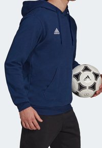 Sweat à capuche bleu marine avec une poche avant et un cordon de serrage, arborant un logo Adidas blanc sur la poitrine. Personne tenant un ballon de football noir et blanc.