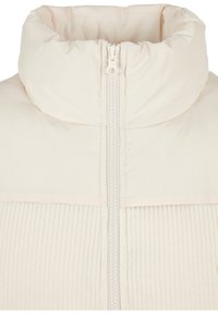 Veste couleur crème avec col haut rembourré, texture côtelée verticale sur la partie inférieure et fermeture éclair centrale.