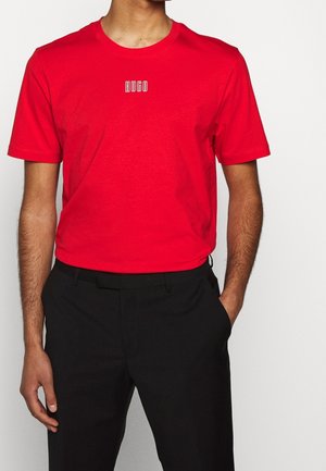 Mann trägt rotes T-Shirt mit Rundhalsausschnitt und weißem "HUGO"-Logo auf der Brust sowie schwarze Hose, eine Hand in der Tasche, vor einfarbigem Hintergrund.